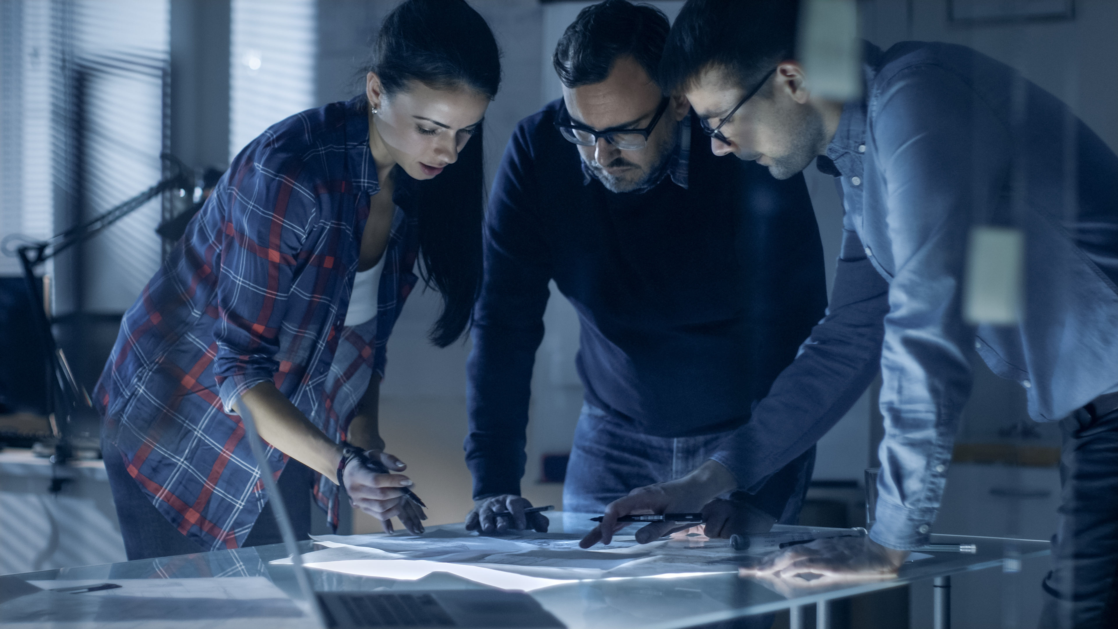 Late at Night Team of Design Engineers Collectively Work on the Project. They Inspect Drafts, Documents and Have Discussion at the Big Table.